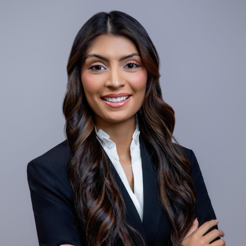 Portrait of Jessica Mendoza, Associate Attorney, smiling with arms crossed. She has long wavy dark hair and is wearing a black blazer with a white shirt, standing against a neutral gray background.
